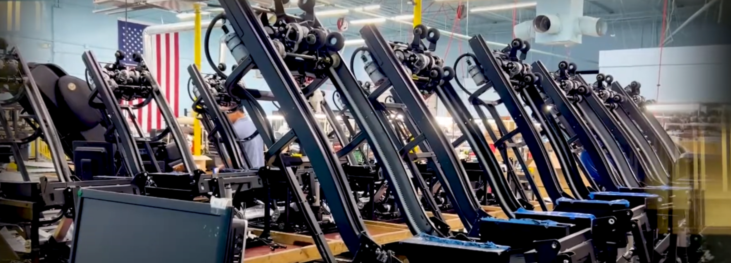 Massage chair frames on the production line with technicians working in a U.S. factory, American flag displayed in the background.