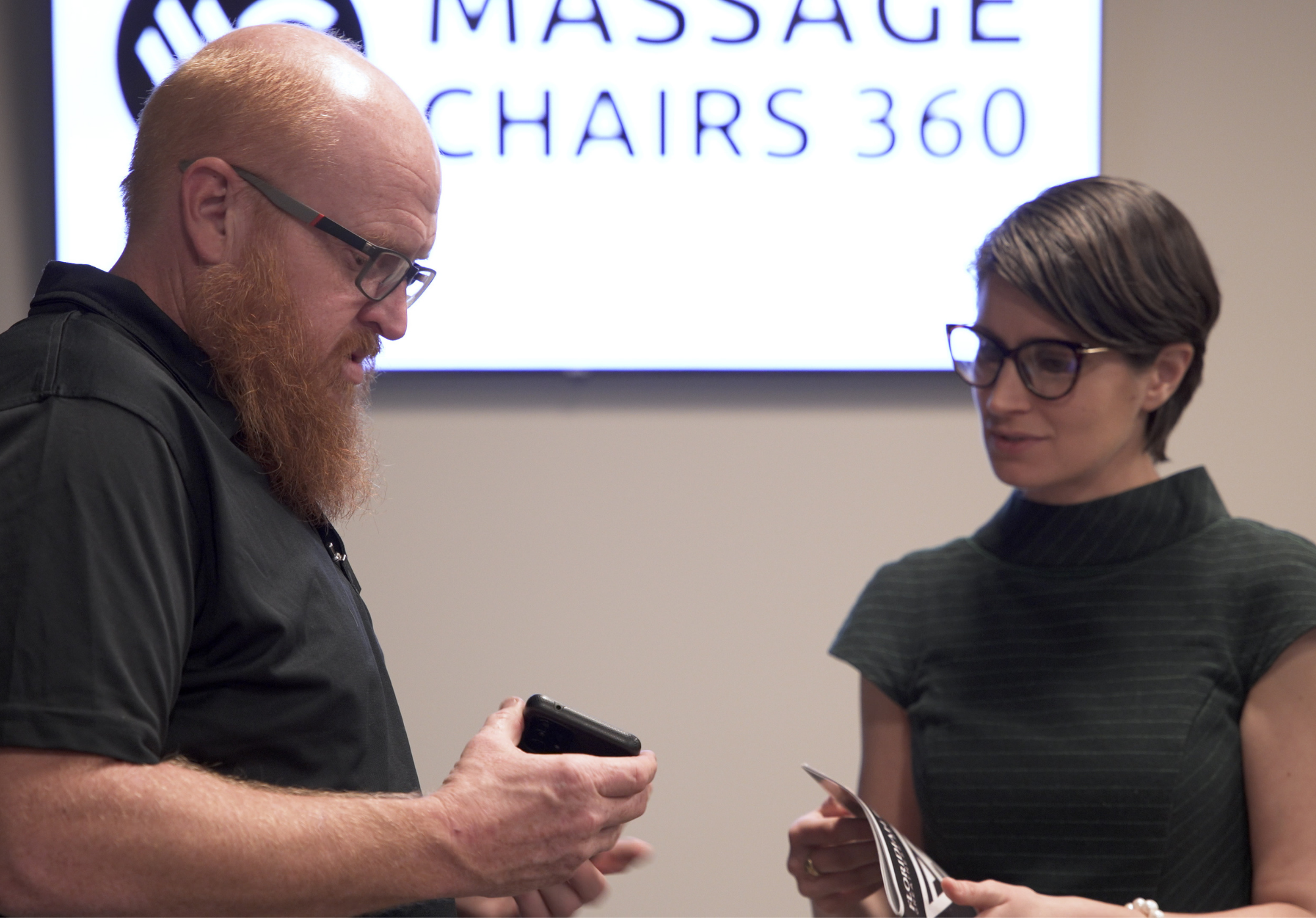 Two people interacting in front of a screen displaying 'Massage Chairs 360'.